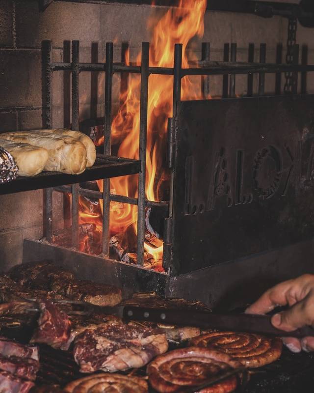 A brasa de perto: fogo alto atrás da chapa de ferro vazada com a marca LALOKA, pães na grelha de cima e carnes e linguiças na de baixo.